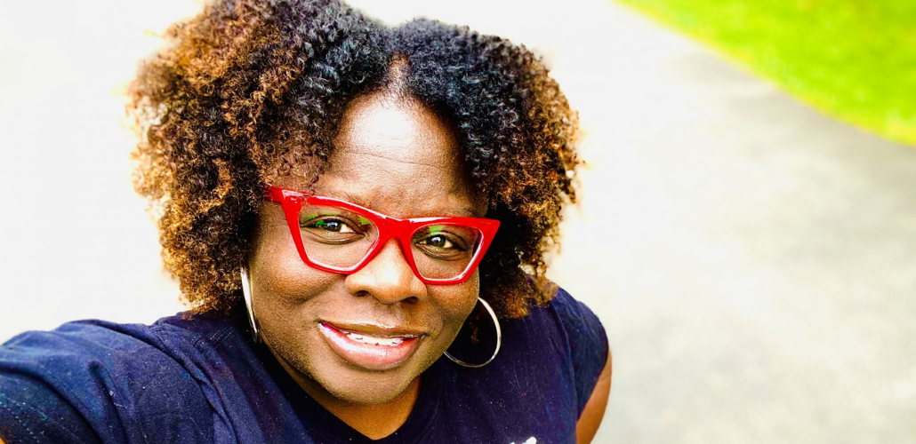 Close-up selfie of Petal Robertson, a Black woman with red glasses, smiling into the camera, outdoors