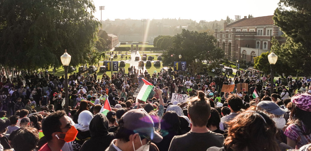 A large crowd, mostly viewed from behind, is arrayed on a hillside or steps of a university campus; banners identify it as UCLA. A few in the crowd hold Palestinian flags and one handmade sign reads "Stay strong." A row of parked vehicles is visible in the distance, at the foot of the hill, backs to the crowd. Dramatic sunlight silhouettes everyone.