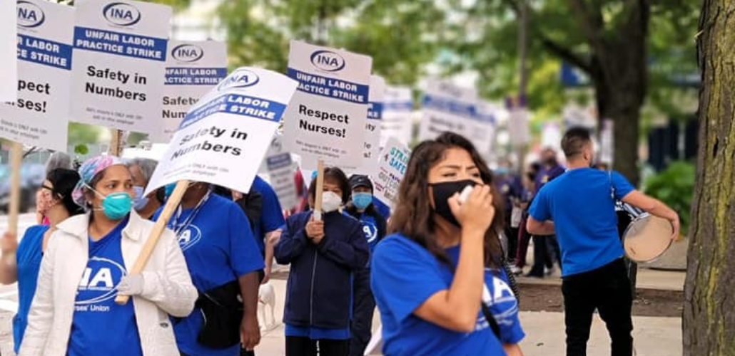 People in blue "INA" shirts march. Some carry picket signs "Safety in Numbers," "Respect Nurses!" "Unfair Labor Practice Strike." One speaks into a bullhorn mic.
