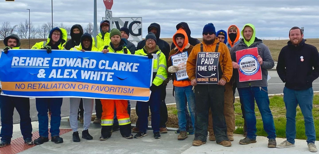16 people stand facing the camera. Most are wearing jeans and neon yellow or orange jackets or hoodies. Several are helping to hold a blue banner: "Rehire Edward Clarke and Alex White, no retaliation or favoritism." Others hold signs: "180 hours paid time off, unionize KCVG" and "Unionize Amazon."