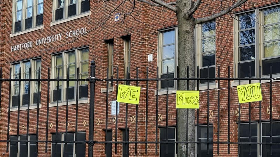 handwritten signs "we miss you" hang on the fence outside a brick building labeled Hartford University School