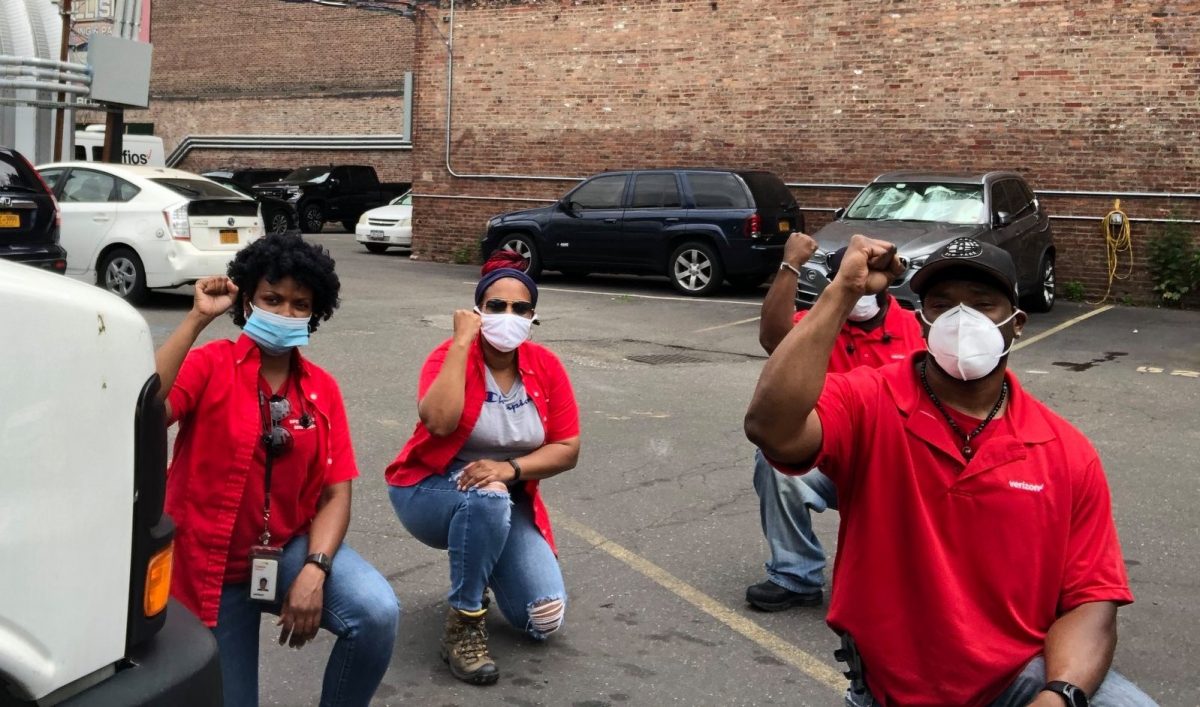 Four Black workers in red Verizon shirts kneel in a parking lot, fists raised.