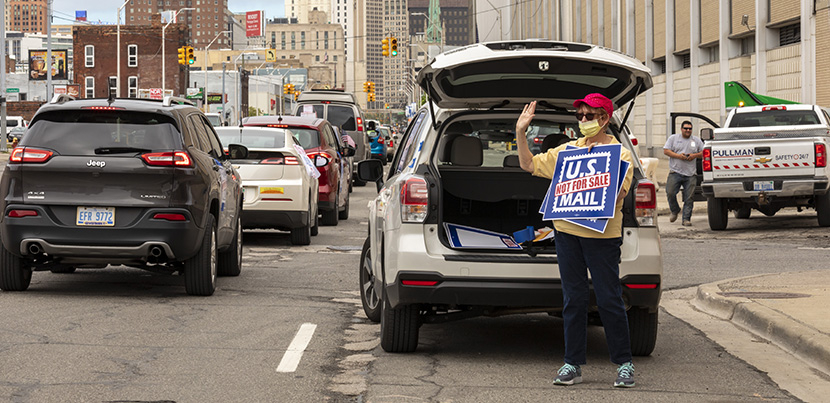 woman in front of the back of her car, trunk open, standing with a save usps sign.