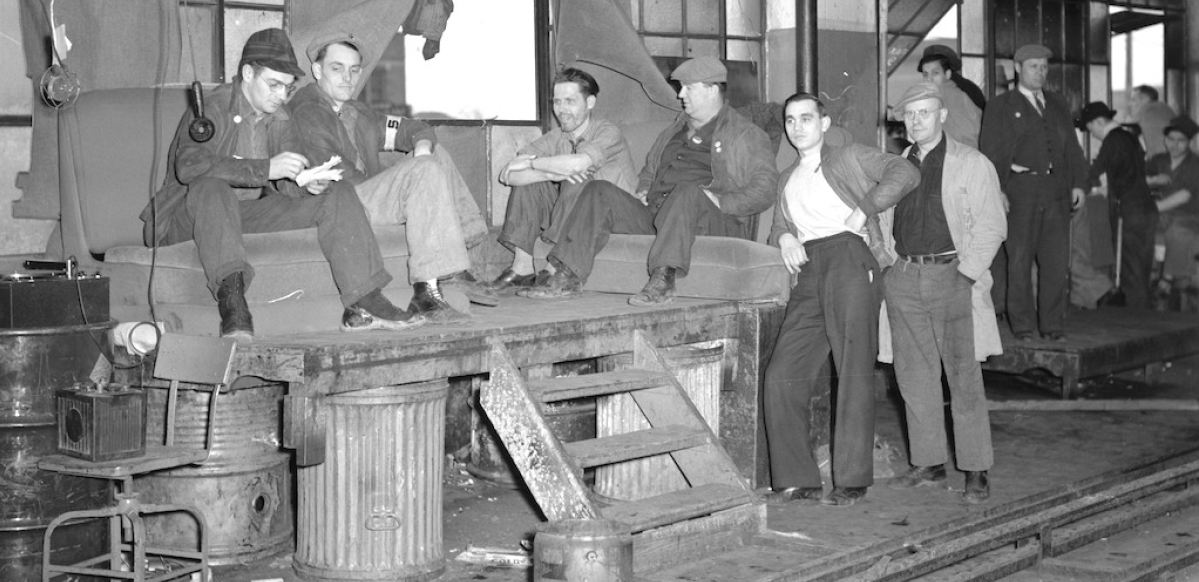 A group of autoworkers sit on a makeshift platform under a window in a factory.