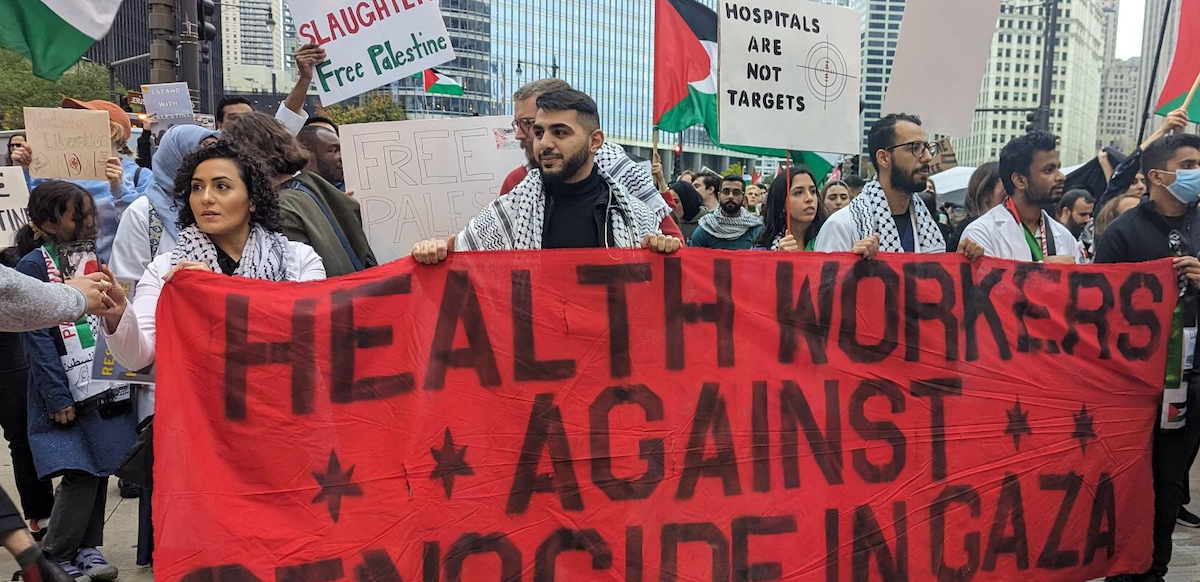 A group of people in white medical coats stand behind a red banner reading ‘Health Workers Against Genocide in Gaza.’