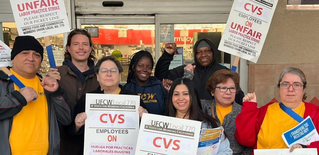 Eight people, varied in race and gender, pose together in front of a CVS store. Some wear UFCW yellow shirts and most carry picket signs that say "UFCW Local 770: CVS employees on unfair labor practices strike, please respect the picket line" in English and Spanish.