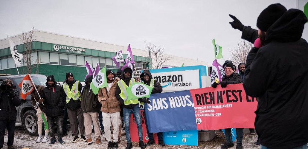 Workers, most of them Black, bundled against the cold, stand in front of an Amazon delivery facility sign. Their banner says, in French, "Without us, nothing is free" and the name of the union. Many hold CSN flags, and smile resolutely. On the right, a figure viewed from back silhouette speaks to them, one hand pointing the way forward.