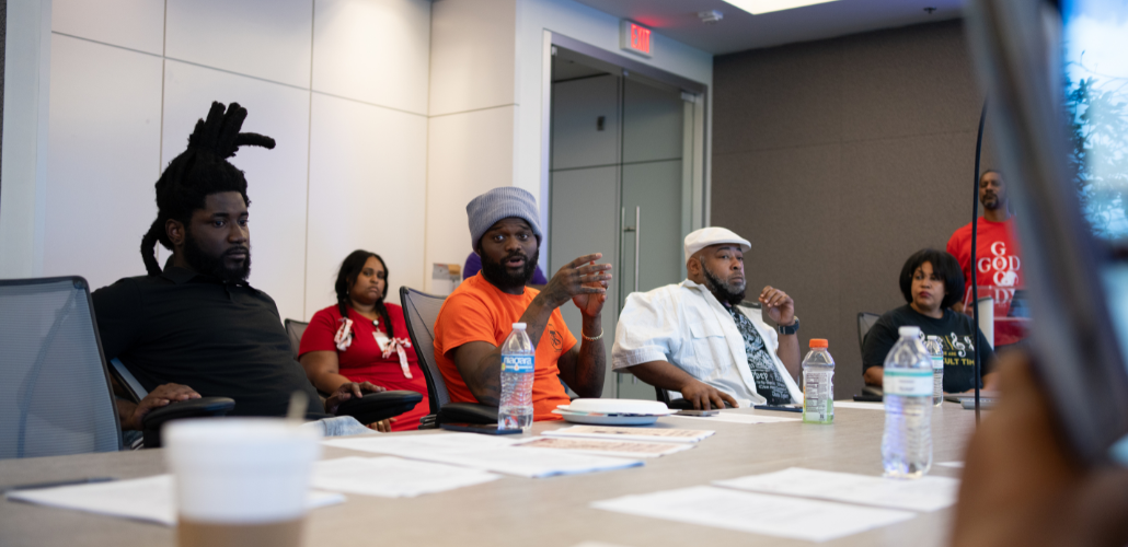 Several Black workers, women and men, sit around a conference table. In the middle of the frame, a man in an orange T-shirt is talking, looking around at the group; others are listening thoughtfully. Everyone's expression is serious.