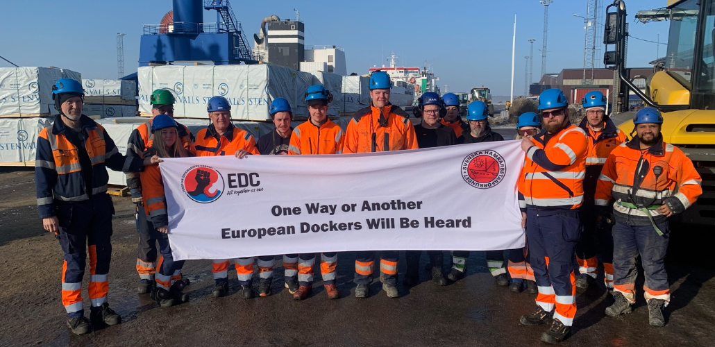 Fourteen workers in orange vests, blue hardhats, and workboots stand in a port in bright sunlight. They hold a big white banner that says "One way or another, European dockers will be heard" with logos of the SDU and the European Dockworkers Council. Most but not all appear to be white men.