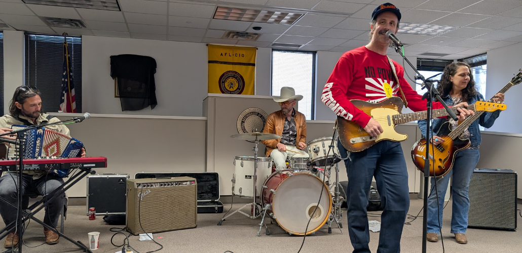A band performs in a union hall. The guitarist in front is a tall man in a red T-shirt with a design that starts "No Power" (his guitar blocks the rest of hte slogan" and the arm says "Stronger." There's a woman on bass, a man in a cowboy hat on drums, and a man holding an accordion sitting at a keyboard. Behind them is a yellow AFL-CIO banner. For some reason a black T-shirt is draped over a framed item on the wall.
