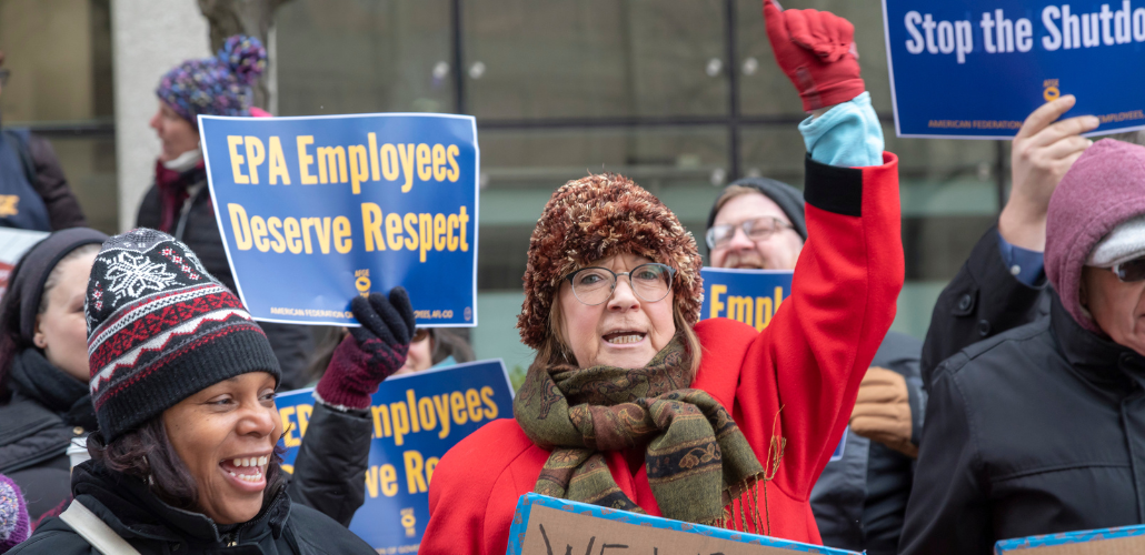 A crowd of federal workers stand outside, bundled against cold, carrying printed AFGE signs that say "EPA employees deserve respect" and "Stop the shutdown." In the foreground are a Black woman and a white woman, the latter raising a red-gloved fist in the air, both chanting or singing.