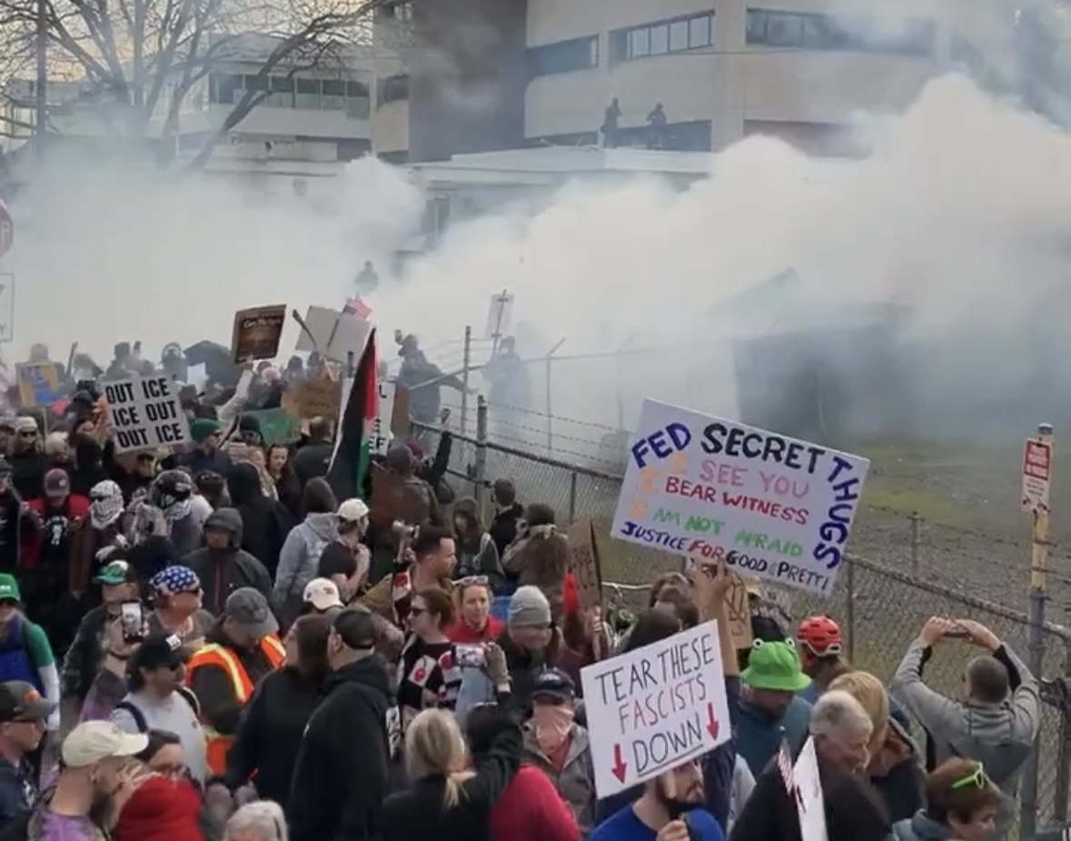 A dense cloud of tear gas envelops a crowd with colorful signs.