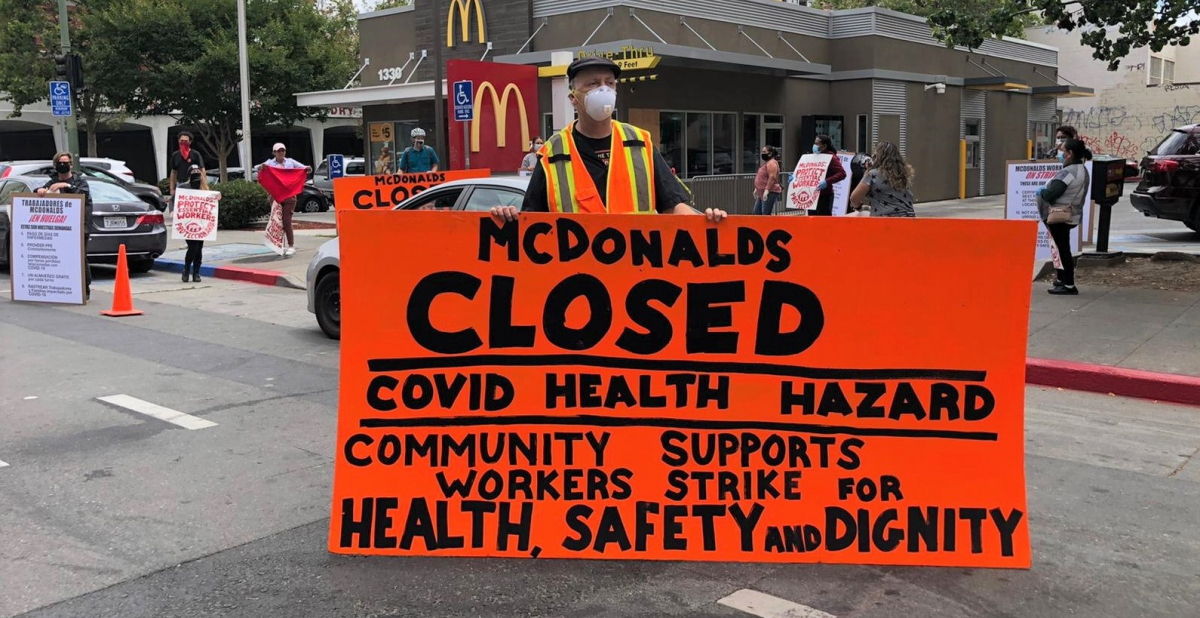 masked worker in front of mcdonald's holds big orange sign: "MCDONALD'S CLOSED, COVID HEALTH HAZARD, COMMUNITY SUPPORTS WORKERS STRIKE FOR HEALTH, SAFETY AND DIGNITY"