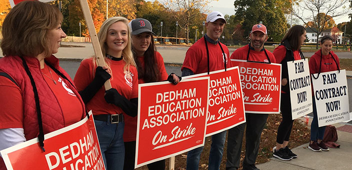 Line of workers on picket line in red union T-shirts holding signs that say "Dedham Education Association on Strike" and "Fair Contract Now" 