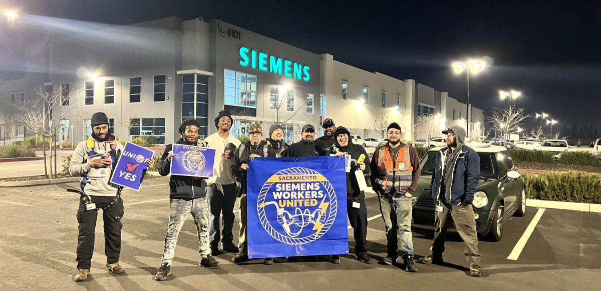 Siemens workers pose in front of the factory in the dark behind a blue banner that says Siemens Workers United.