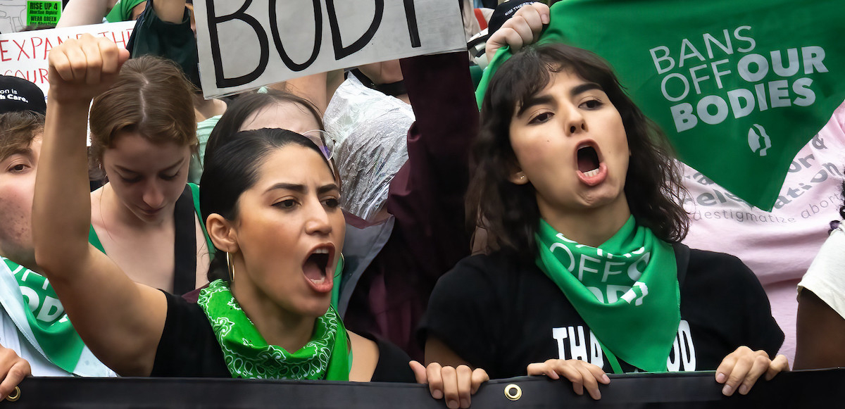 Two women in green bandanas that say “bans off our bodies” march with many others