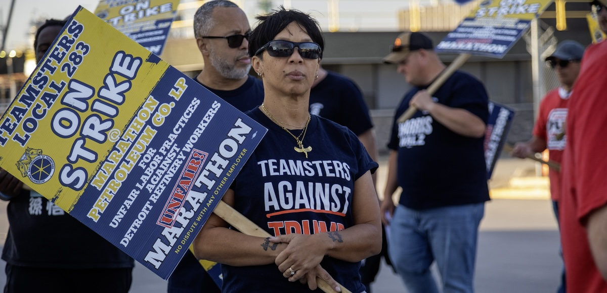 A woman with a strike picket sign and a ‘Teamsters Against Trump’ T-shirt looks past the camera