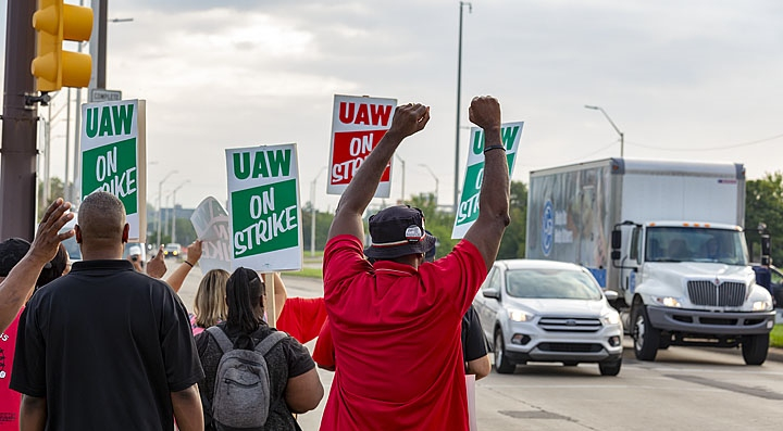 Strikers shown from behind, waving at cars, fists in air, signs read "UAW On Strike"