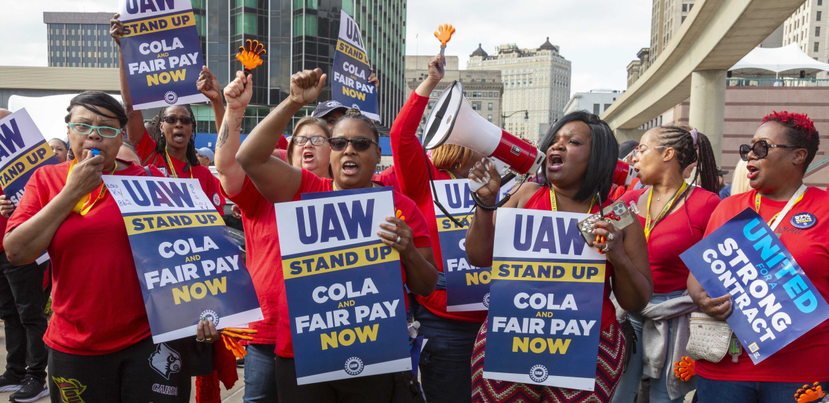 Workers in red shirts chant. Most are Black women. Many fists in air, one blowing whistle, one speaking into bullhorn. Printed signs say "UAW Stand Up, COLA and Fair Pay Now" 