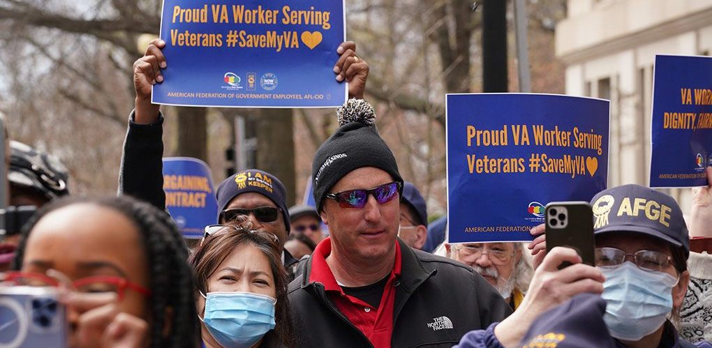 several people are jammed together as part of a march with blue signs with gold lettering, saying “Proud VA Worker Serving Veterans, #SaveMyVA”