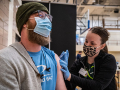 Woman on right in mask administering covid vaccine to man in mask and hat on left.