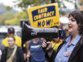 A woman with long dark hair speaks into a megaphone to a crowd that is visible beyond her, one holding a “Fair Contract Now” sign.