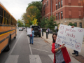 A teacher holds a sign between a school bus and the front of a school, with students visible in the background