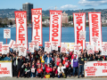 A crowd of hundreds is seen from afar and behind them there’s a body of water. They have vertical red on white banners saying “Health and Safety,” “Racial Justice,” “For the schools,” “That Students,” “Deserve.” 