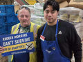 Two Kroger workers stand in front of pallets. One holds a sign that says "Kroger workers can't survive off of quarters."