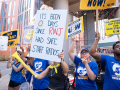 Nurses in blue T-shirts blow whistles and air horns outside a red-brick building with huge white columns.