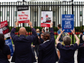 A line of seven flight attendants in uniform, viewed from behind, stands at the White House gates holding up various printed signs. Messages say: "AA makes billions, we can't pay rent." "Robert, what's the delay? We need a contract today!" "American Airlines flight attendants: No retro pay, no way!" "Flight attendants save lives!" "Corporate greed doesn't fly." "Trying to get pAAid." It appears the group includes men and women, and most are people of color.