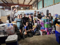 A group of fifteen workers pose inside a warehouse among boxes of diapers and other aid material