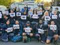 At least 32 letter carriers in uniform, holding "no" signs printed in various fonts, pose in a post office parking lot. The group looks diverse in race and gender. Several give the "thumbs-down" hand signal.