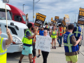 Workers in high-visibility vests picket with ‘Amazon workers on strike’ signs picket by a road, one handmade sign reads ‘Stop the captive audience’