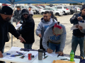 A group of men in sweatshirts gather around a table and sign things, a parking lot is behind them.