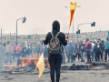 Protester holding what appears to be a flag and a torch in front of burnt out remains. Small crowd in background.