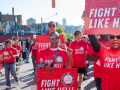 Black and white women and men march through a Detroit street. Everyone is wearing a red "fight like hell" T-shirt with the NALC logo, and some people are carrying printed signs with the same design. Most prominent in the frame, looking directly into the camera, are two Black women in sunglasses, one raising a fist at shoulder level and the other holding a sign above her head. 