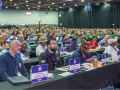 UAW delegates (men and women, Black and white) sit listening at the union's 2026 convention. Table signs identify those in the foreground as from Region 8, Locals 129 and 276.