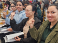Four women of a range of ages, seated in a row, part of a bigger crowd in what looks like a hotel ballroom, smile and raise fists. Each holds in her lap a stapled packet of printed pages, perhaps a summary of a tentative agreement.