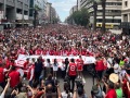 A huge crowd, as far as the eye can see, many wearking red, march down a street flanked by tall buildings.