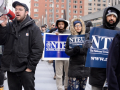 A line of men and women picketers with NTEU signs march on the right while a man with a bullhorn chants on the right