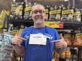 A man in in front of grocery shelves points to a printed-out “Vote No” sign pinned to his blue shop apron.