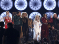 A row of musicians with large clocks behind them perform, with Dolly Parton in the center