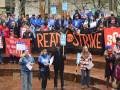 People rally on brick steps (Portland's Pioneer Courthouse Squre) on a rainy day. Many wear blue or red T-shirts under their coats. Banners say "Ready to strike" and "Soldarity." Picket signs say "Fair Contract Now," "Solidarity with Strike-Ready Teachers," and "Essential Wages for Essential Workers."