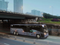 An electric coach bus drives under an underpass with city buildings in the background