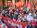 View of hundreds of people, a section of a giant march, filling an Italian street. Some marchers carry a huge red banner "FIOM-CGIL Bologna," some carry union flags or Palestinian flags, and one carries a handmade sign that says "Palestina Libera" with the flag and hearts. Mouths are open to sing or chant. Picturesque columns along the street. Marchers are diverse in race and gender.