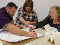 Three people reach across a table to mark a drawing of their workplace