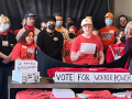 A group of serious-faced people stands indoors. Some are wearing matching red EW4D T-shirts, including Iris Scott, a white woman speaking from notes while others listen and someone in front of them holds up a phone camera to livestream the event. Some in the group wear other union shirts. Handwritten signs on the table in front of the group say "We need true reprsentation," "Vote for worker power," and "UFCW 3000, UAW, TDU" [with hand-drawn logos]. 