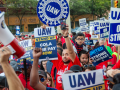 UAW members and supporters at a rally. Many hold signs saying UAW on Strike. Several have their children on their shoulders.