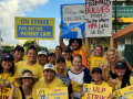 Nurses in yellow shirts pose smiling. Most are women; most appear Native Hawaiian or Asian. Many wear ballcaps. Some carry printed signs saying "ULP strike," "Zero tolerance for retaliation," or "on strike for better patient care." Two carry handmade signs--one with an image of a duck and the words "Unsafe staffing leads to quack-cidents," and the other with the logo of the show "Friends" crossed out, the word "Bullies" written in the same style, followed by "Episode 2: The One Where HPH Locks Us Out."